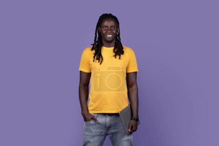 Téléchargez les photos : Positive cool handsome young african american long-haired guy in casual holding modern laptop on purple studio background, black man freelancer posing with modern computer, copy space - en image libre de droit