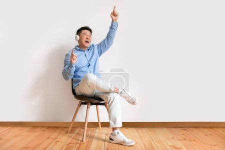 Téléchargez les photos : Joyful asian mature man in wireless headphones sitting on chair and listening to music, raising his arm, dancing to favorite song. Middle aged male enjoying cool soundtrack against white wall - en image libre de droit