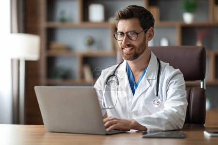 Smiling Handsome Male Doctor Working On Laptop While Sitting At Desk In Office, Happy Physician Man Wearing Medical Coat Consulting Patients Online, Typing On Computer Keyboard, Copy Space