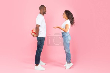 Photo for African american middle aged man hiding gift box behind back and looking at happy interested woman, isolated on pink background, studio shot, side view - Royalty Free Image