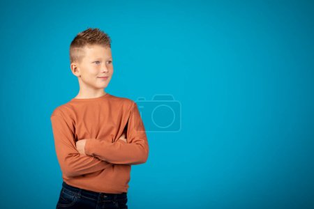 Foto de Portrait Of Handsome Preteen Boy With Folded Arms Standing Over Blue Background, Pensive Male Child Looking Away And Smiling, Thinking About Something While Posing In Studio, Copy Space - Imagen libre de derechos