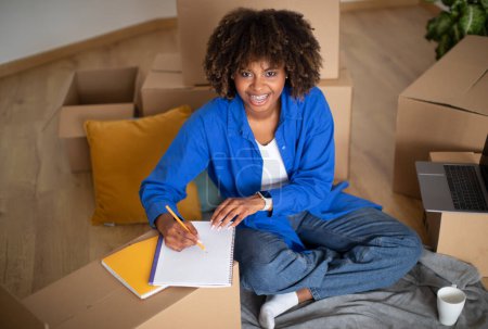 Photo for Happy Young Black Woman Sitting Among Cardboard Boxes After Moving And Writing In Notepad, Smiling African American Female Doing Checklist Of Necessities During Relocation To New Apartment - Royalty Free Image