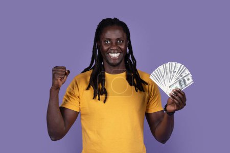 Photo for Emotional happy young african american guy in yellow showing bunch of cash dollars and gesturing, clenching fist, celebrating success over purple studio background. Gambling, trading, giveaway - Royalty Free Image