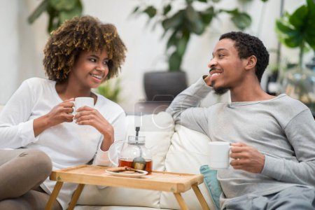 Photo for Cheerful smiling african american young husband and wife sitting on couch in cozy living room, drinking tea, eating cookies, have conversation, spending time together at home. Marriage, relationships - Royalty Free Image