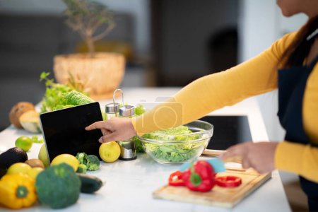 Foto de Young african american woman in apron cuts salad, use tablet with empty screen at table with vegetables in modern kitchen interior, cropped. App for diet, cooking healthy eat at home, food blog lesson - Imagen libre de derechos