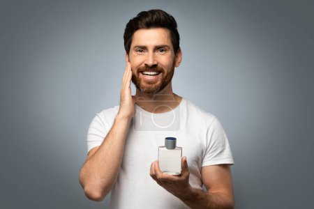 Photo for Grooming and people concept. Happy handsome caucasian man applying perfume on beard, posing over grey studio background and smiling at camera - Royalty Free Image