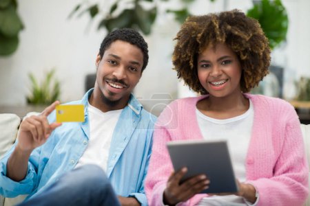 Foto de E-commerce concept. Happy smiling beautiful black couple shopping on Internet from home, sitting on couch in cozy living room, using modern digital tablet and credit card, copy space - Imagen libre de derechos