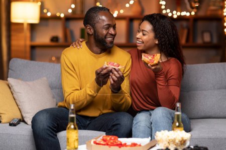 Téléchargez les photos : Portrait of happy african american lovers man and woman sitting on couch in living room decorated with Christmas lights, eating pizza, drinking beer, laughing and hugging at home - en image libre de droit