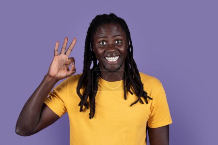 Téléchargez les photos : Cool cheerful handsome young black man in casual outfit with long braids showing okay, smiling on purple studio background, like something or someone, copy space. Human gestures concept - en image libre de droit