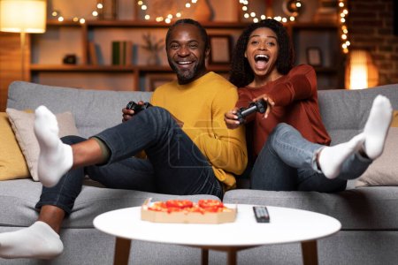 Téléchargez les photos : Emotional happy black spouses sitting on couch, holding joysticks and laughong, couple playing video games at home, eating pizza, living room decorated with festive lights, copy space - en image libre de droit