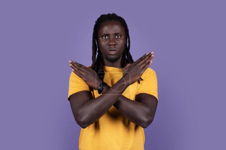 Foto de Serious african american millennial guy in casual saying no, looking at camera and showing hands crossed over chest, purple studio background, copy space. Human gestures, cross sign concept - Imagen libre de derechos