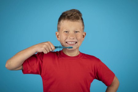 Téléchargez les photos : Happy Preteen Boy Brushing Teeth With Toothbrush And Smiling At Camera, Cheerful Caucasian Male Child Making Daily Dental Care, Standing Isolated Over Blue Studio Background, Copy Space - en image libre de droit