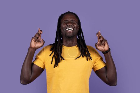 Téléchargez les photos : Making Wish. Superstitious emotional stylish black guy in yellow t-shirt with closed eyes keeping his fingers crossed, praying for luck, isolated on purple studio background, copy space - en image libre de droit