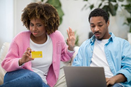 Foto de Annoyed pretty curly young black woman holding credit card while her husband or boyfriend using laptop, checking prices on webstore or bank account on Internet, couple banking online from home - Imagen libre de derechos