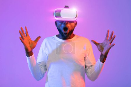Foto de Augmented reality, modern technologies concept. Shocked emotional african american bearded guy in white with shining wireless VR headset gesturing, raising hands up, neon light studio background - Imagen libre de derechos