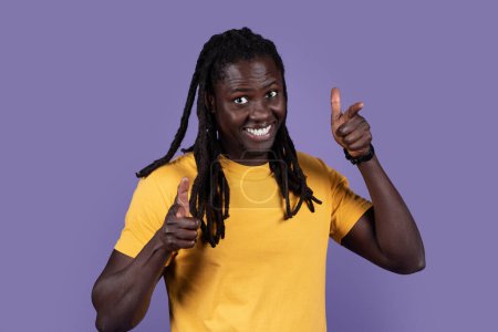 Photo for Hey you. Charismatic cheerful handsome young long-haired black man in yellow t-shirt pointing at camera with both hands, flirting over purple studio background, making choice, copy space - Royalty Free Image