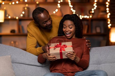 Téléchargez les photos : Handsome african american man greeting his beautiful wife with St. Valentines Day, cheerful black husband giving his excited woman gift box and hugging her, festive home interior, copy space - en image libre de droit