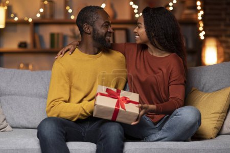 Foto de Loving beautiful young african american woman greeting her husband with St. Valentines Day, cheerful black lady giving boyfriend gift box and hugging him, festive home interior, copy space - Imagen libre de derechos