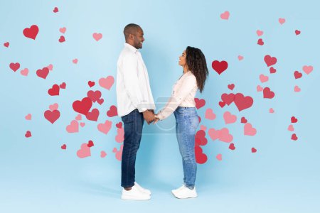 Photo for Full length shot of happy black couple holding hands and looking at each other on blue studio background with red flying hearts, side view. St. Valentines Day - Royalty Free Image