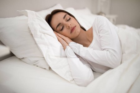 Foto de Healthy sleeping concept. Peaceful young attractive brunette woman wearing white long sleeve pajamas sleeping in comfy bed on soft pillows with hands under her face at home, copy space - Imagen libre de derechos