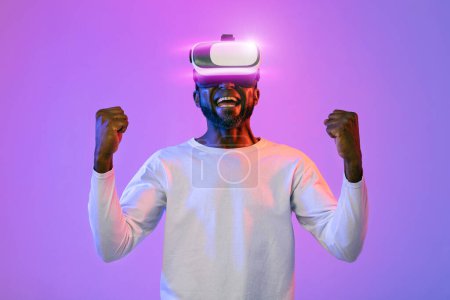 Photo for Emotional happy bearded african american man in white trying virtual reality headset, clenching fists and screaming over neon light studio background. Modern technologies and entertainment concept - Royalty Free Image