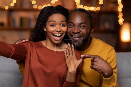 Photo for Happy emotional beautiful black couple got engaged on Valentines evening, cheerful man and woman sitting on couch at festive home interior, taking selfie together, showing engagement ring and smiling - Royalty Free Image