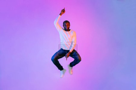 Téléchargez les photos : Funny cheerful smiling middle aged handsome african american man in casual jumping in the air and gesturing, pointing up and down, neon light studio background, full length, copy space - en image libre de droit