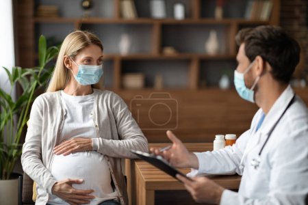 Photo for Young Pregnant Female Wearing Face Mask Having Check Up With Therapist Doctor, Gynecologist Man Consulting Expectant Woman During Meeting In Hospital, Prescribing Vitamins Or Pills, Free Space - Royalty Free Image