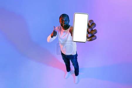 Photo for Cheerful middle aged black man showing big cell phone with white blank screen and thumb up, recommending nice online offer, mockup, copy space, neon studio background, full length, high angle view - Royalty Free Image