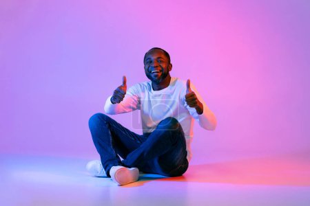 Téléchargez les photos : Cool happy handsome middle aged black man in casual outfit sitting on floor, showing thumbs up and smiling on colorful neon studio background, copy space for advertisement, full length - en image libre de droit