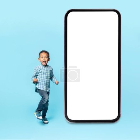 Foto de Emotional black kid boy clenching fists, standing near big smartphone with white blank screen, mockup, blue studio background, cropped - Imagen libre de derechos