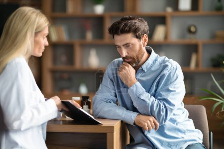 Foto de Female Physician Discussing Check Up Results With Worried Man During Meeting At Hospital, Thoughtful Male Patient Touching Chin While Doctor Lady Explaining Him Details Of Treatment, Closeup - Imagen libre de derechos