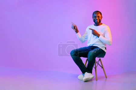 Téléchargez les photos : Cheerful happy handsome african american middle aged man in casual outfit sitting on chair, pointing at free space for advertisement with both hands over neon studio background, full length - en image libre de droit
