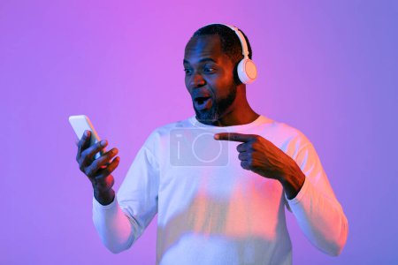 Foto de Amazed happy handsome middle aged african american guy with wireless headphones poitning at smartphone in his hand, black man using exciting entertaining mobile app on phone over neon light background - Imagen libre de derechos