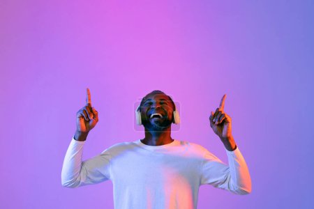 Photo for Happy emotional cheerful african american handsome bearded middle aged man enjoying his brand new wireless stereo headphones, raising fingers up at copy space for ad above and smiling - Royalty Free Image