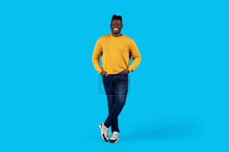 Téléchargez les photos : Handsome black man standing with hands in pockets while posing over blue background in studio, happy young african american man in casual clothes smiling at camera, full length shot, copy space - en image libre de droit