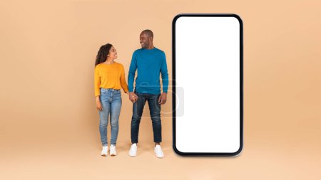 Foto de Loving black couple standing near large smartphone with blank screen, holding hands and looking at each other, advertising offer or mobile app, mockup, full length shot - Imagen libre de derechos
