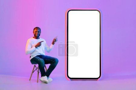 Téléchargez les photos : Happy smiling middle aged black man sitting on chair by big cell phone, pointing at white blank smartphone screen, showing nice online offer or mobile app, mockup, neon light studio background - en image libre de droit