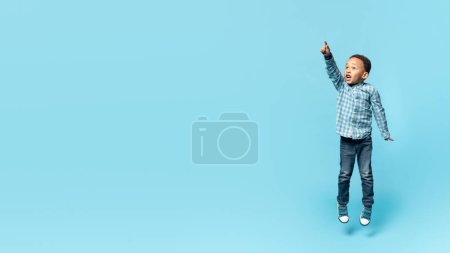 Foto de Emotional little african american kid boy jumping up and pointing at copy space for advertisement over blue studio background, showing something exciting, panorama, full length - Imagen libre de derechos