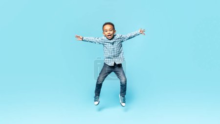 Téléchargez les photos : Happy little african american boy posing in mid air, jumping and spreading arms over blue studio background, full length. Carefree child having fun. Kids fashion and style - en image libre de droit