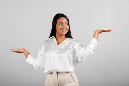 Foto de Glad confident millennial black female spreads her arms to sides, hold copy space, choice ad and offer, isolated on gray background, studio. Comparison of business, work, investment, study and startup - Imagen libre de derechos