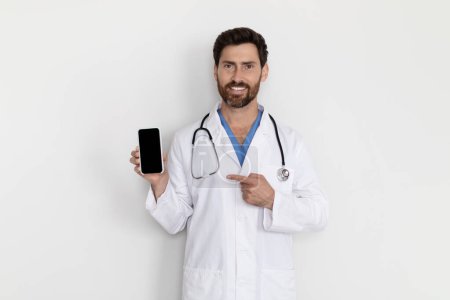 Téléchargez les photos : Medical Insurance. Handsome Male Doctor Wearing Uniform Pointing At Blank Smartphone In His Hand While Standing Near White Wall In Hospital, Therapist Man Recommending Online Offer, Mockup - en image libre de droit