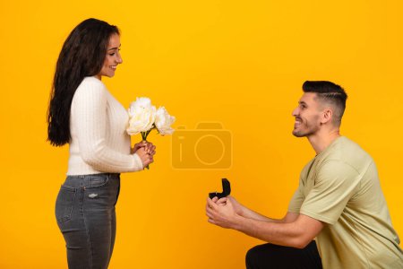 Téléchargez les photos : Glad millennial arabic male on knees give box with ring to smiling lady with bouquet of flowers, isolated on orange background. Engagement, love, relationship and romance, surprise marriage proposal - en image libre de droit