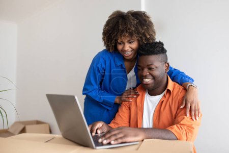 Photo for Young Happy Black Couple Using Laptop Computer In Their New Home After Moving, African American Spouses Making Online Shopping, Ordering Furniture In Internet While Sitting Among Cardboard Boxes - Royalty Free Image