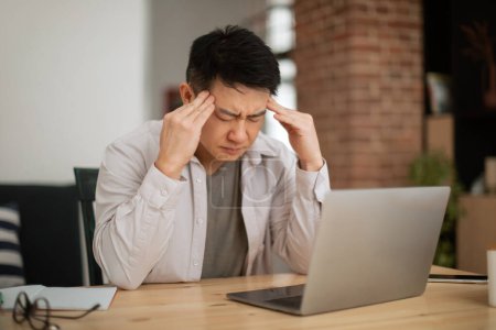 Photo for Sad mature asian male freelancer working on laptop and suffering from headache, sitting at table at home office, copy space. Upset male looking at computer screen, reading bad news online - Royalty Free Image
