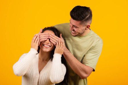 Téléchargez les photos : Smiling millennial arabic guy closes eyes of surprised lady, birthday greeting, isolated on orange background, studio. Celebrate anniversary and surprise for Valentine day, family love and romance - en image libre de droit