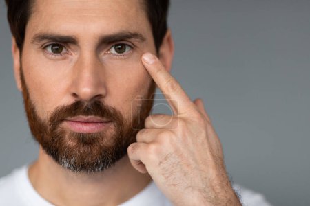 Foto de Male facial skincare. Handsome caucasian bearded man touching face, caring for skin under eyes, looking at camera, standing over grey studio background. Closeup shot, free space - Imagen libre de derechos