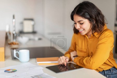 Photo for Smiling Young Arab Female Browsing Internet On Digital Tablet In Kitchen, Beautiful Young Middle Eastern Woman Using Modern Gadget For Online Work Or Study At Home, Closeup Shot With Free Space - Royalty Free Image