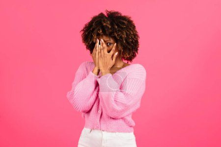 Foto de Frightened millennial african american curly woman in casual wear covering face with hands and afraid, isolated on pink background, studio. Human emotions, fear, reaction to bad news and horror film - Imagen libre de derechos