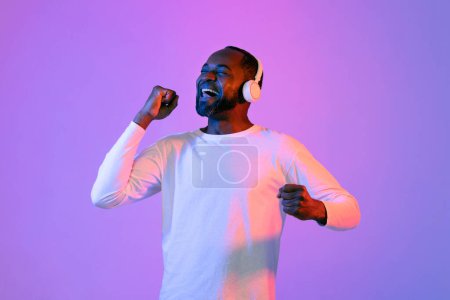 Foto de Cheerful middle aged handsome bearded black man in white listening to favorite music and dancing over neon light studio background, using modern wireless headphones, copy space - Imagen libre de derechos